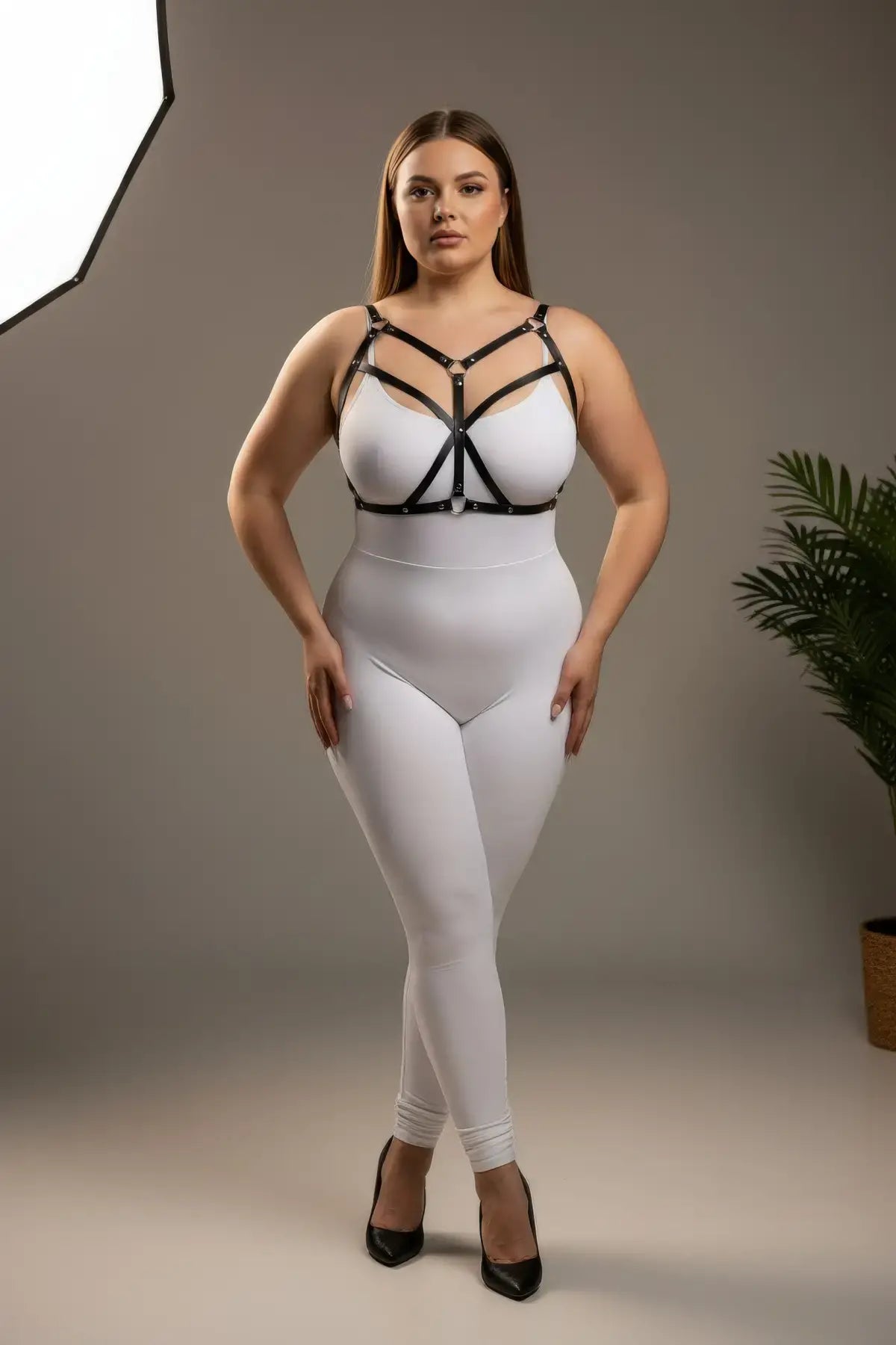Plus-size black cross-strap faux leather bustier harness by HarnessClub, worn over a white bodysuit.