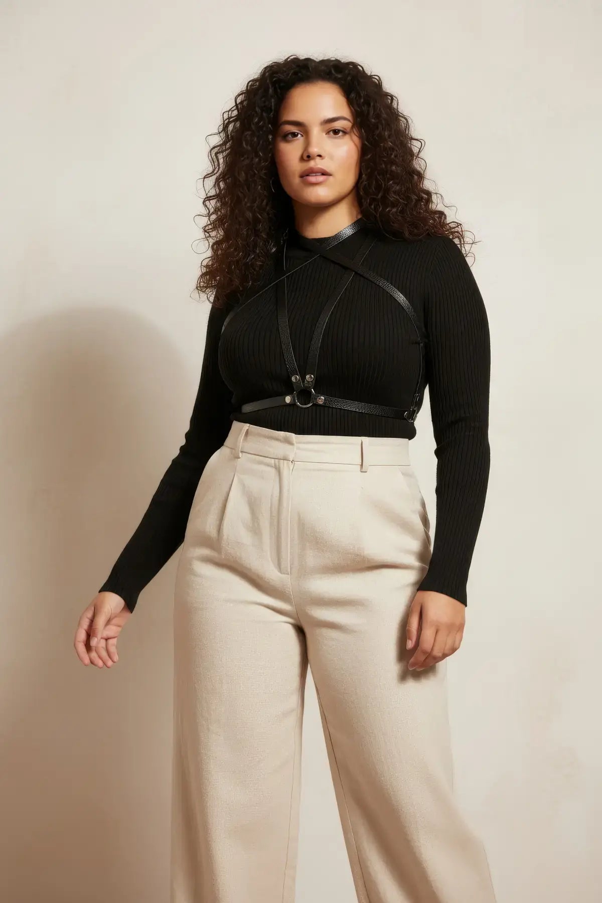 Plus-size black vegan-leather crossing-strap cage harness worn over black rib-knit top and beige trousers