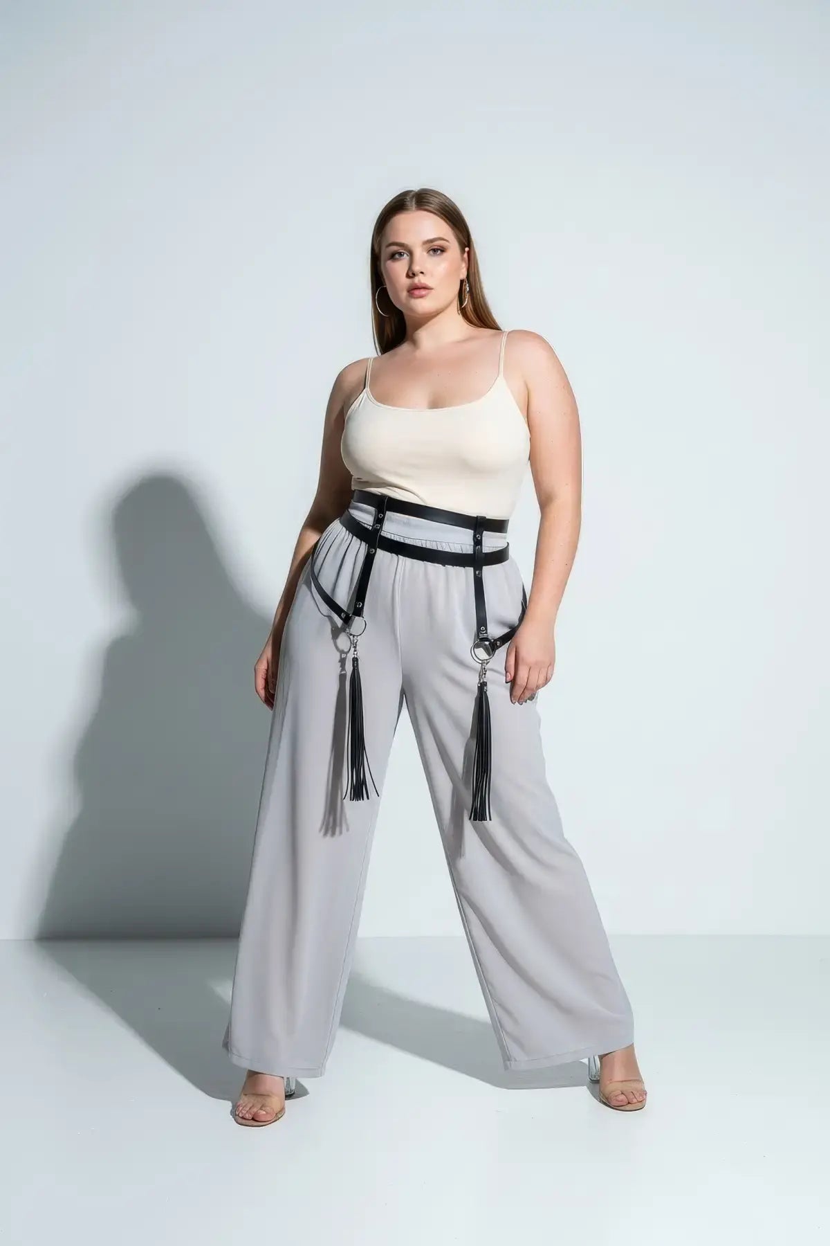 Plus-size black garter hip belt with leg-strap tassels by HarnessClub, shown on a model in a cream top and gray pants.