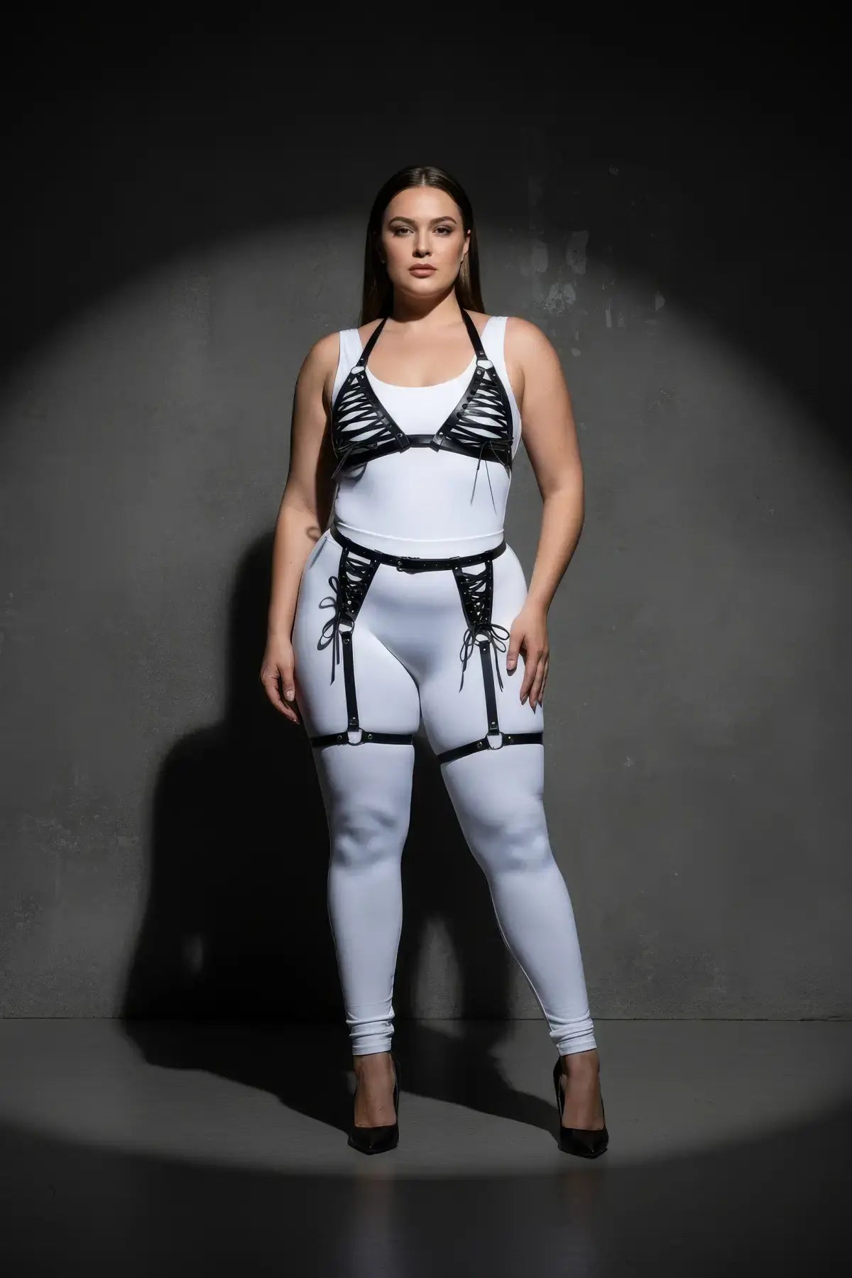 Plus-size black vegan-leather lace-up harness and garter belt set worn over a white bodysuit.
