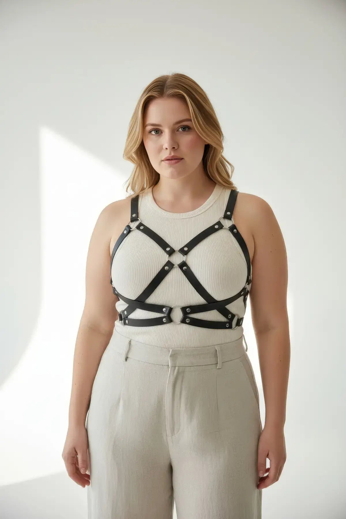 Plus-size black leather harness with O-ring hardware worn over a beige ribbed top