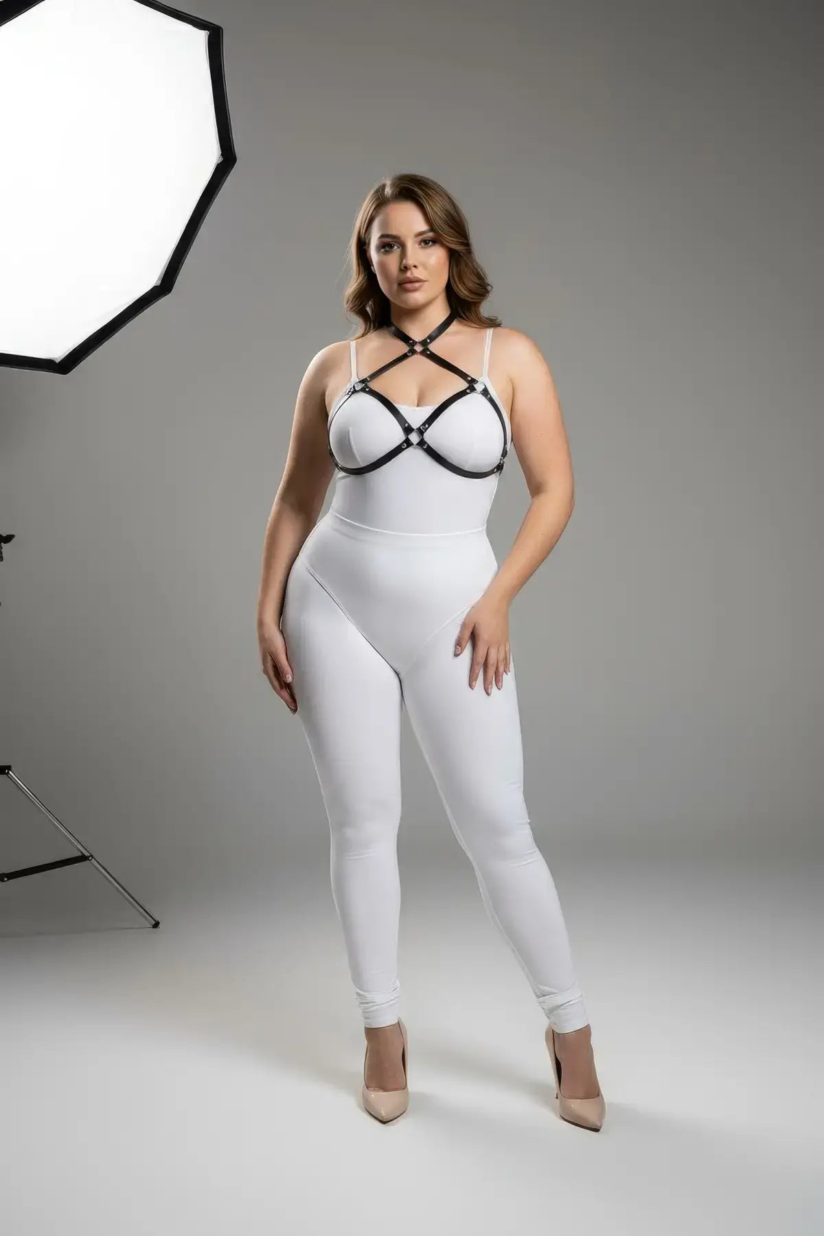 Plus-size black faux-leather O-ring chest harness with metal hardware, worn over a white bodysuit and beige heels.