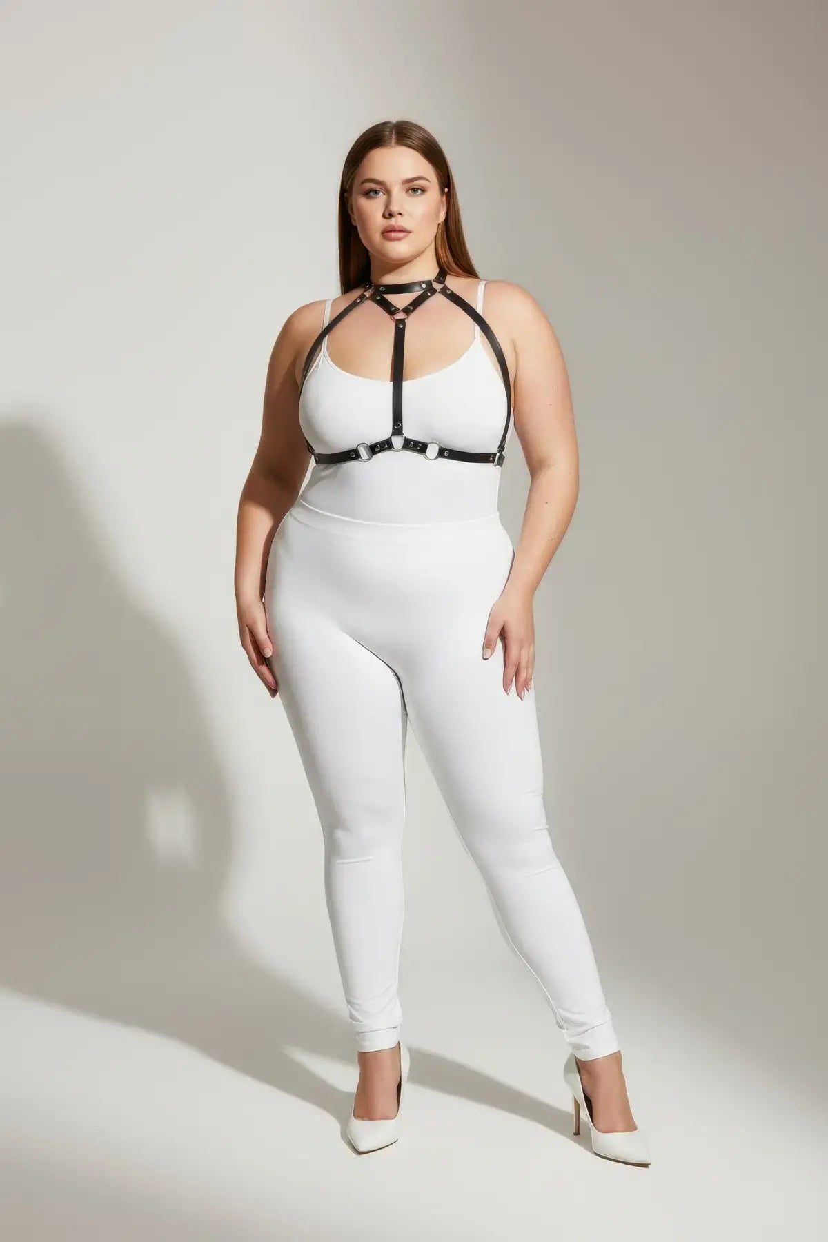 Plus-size black faux-leather O-ring harness worn over a white bodysuit; adjustable straps and rings by HarnessClub.
