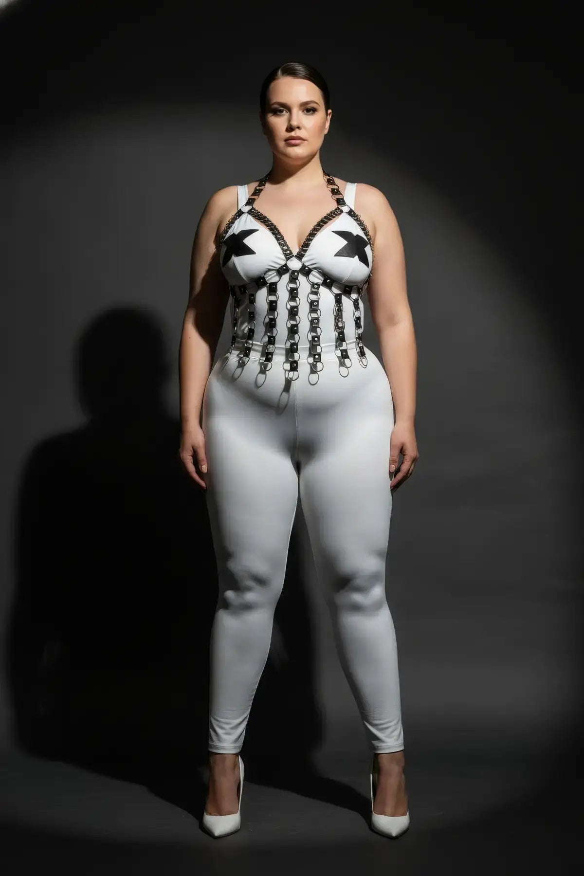 Plus-size black vegan-leather chest harness with studded V-frame and O-ring fringe on white bodysuit.