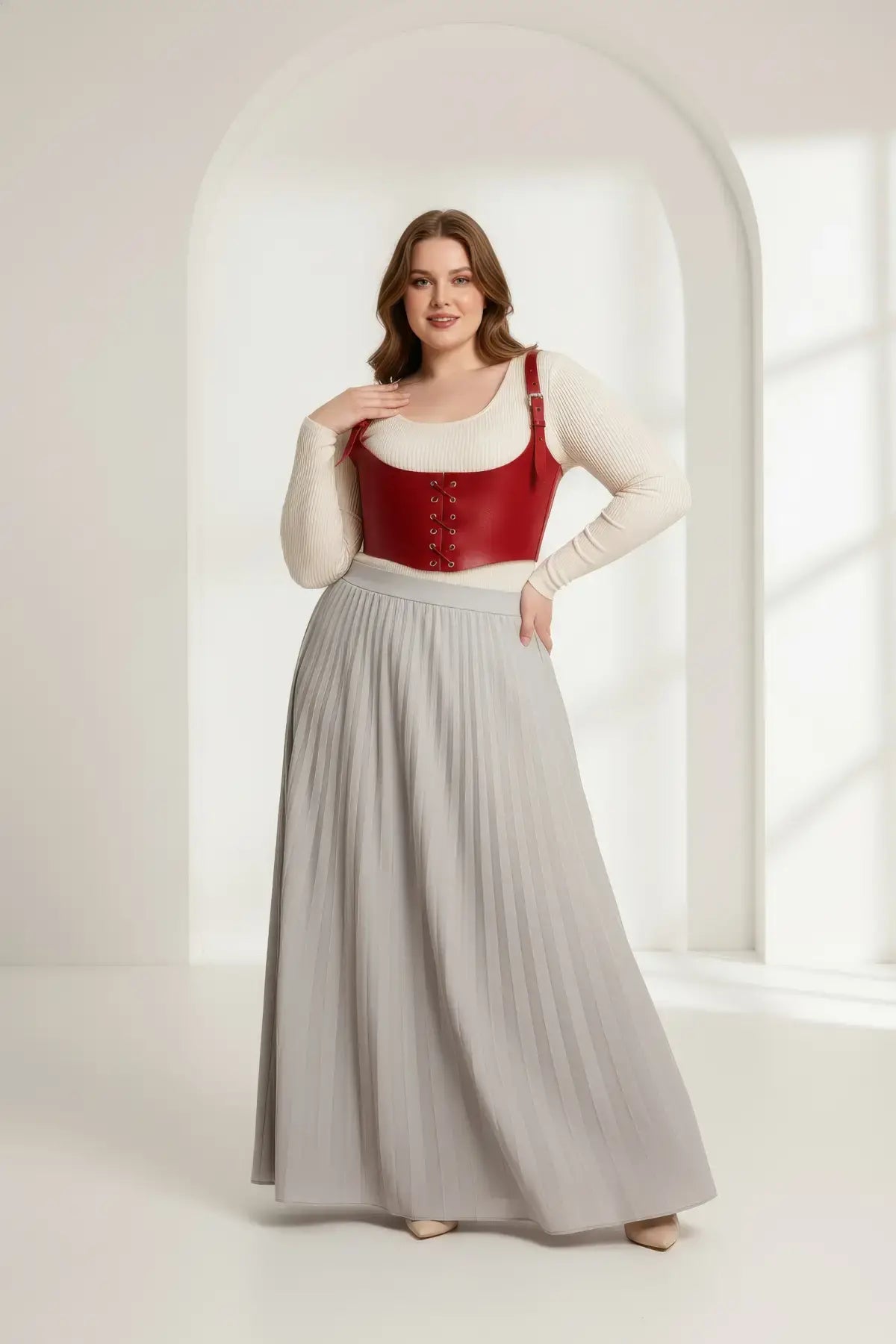 Plus-size red vegan leather lace-up corset harness worn over a cream knit top with a gray pleated maxi skirt