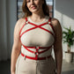 Plus-size red leather corset-detail body harness with cross straps and metal rings worn over a cream ribbed tank top