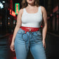 HarnessClub plus-size red vegan leather belt with silver eyelets, O-ring and chain detail worn over jeans.