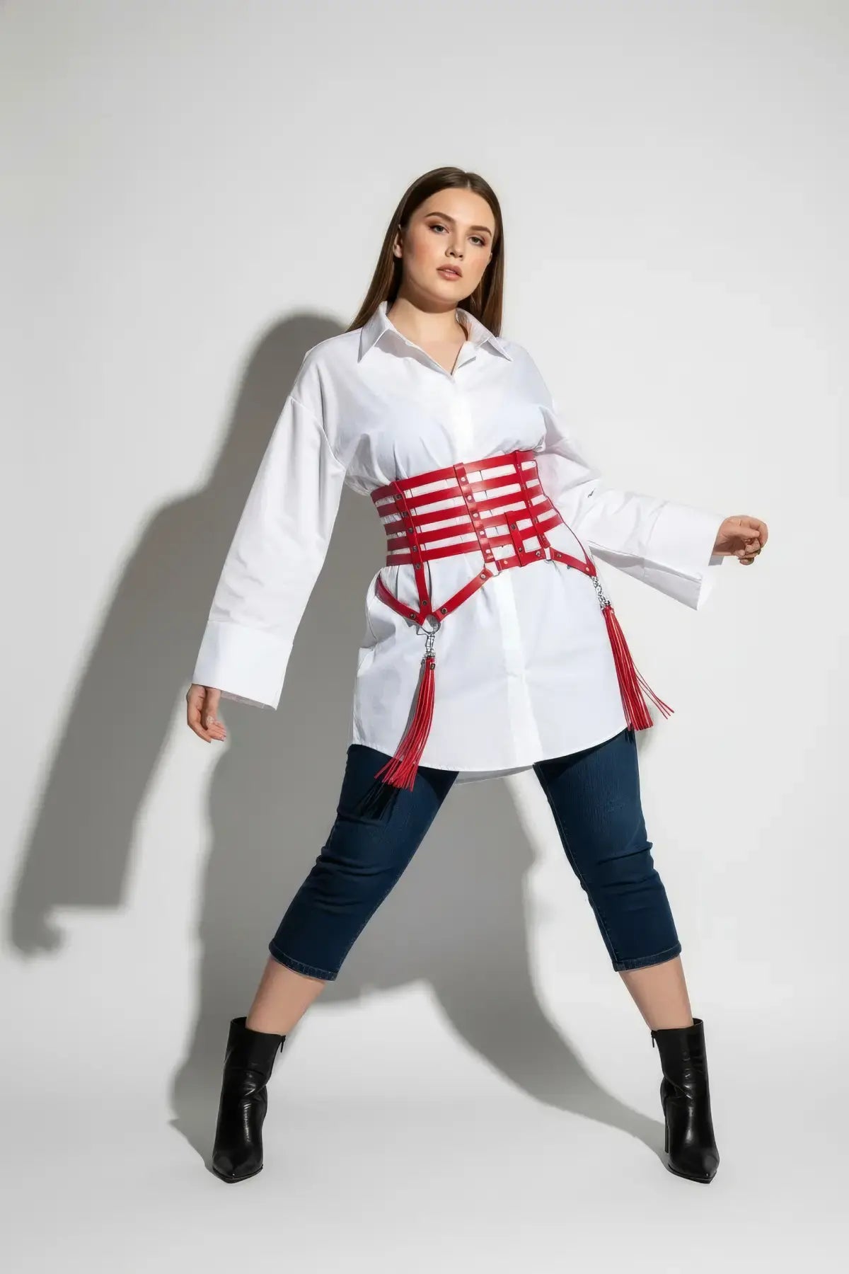 Plus-size red vegan leather corset belt with six horizontal straps and tassels, worn over a white shirt dress.