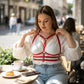 Plus-size red vegan-leather shoulder-strap corset harness worn over a white blouse at an outdoor cafe