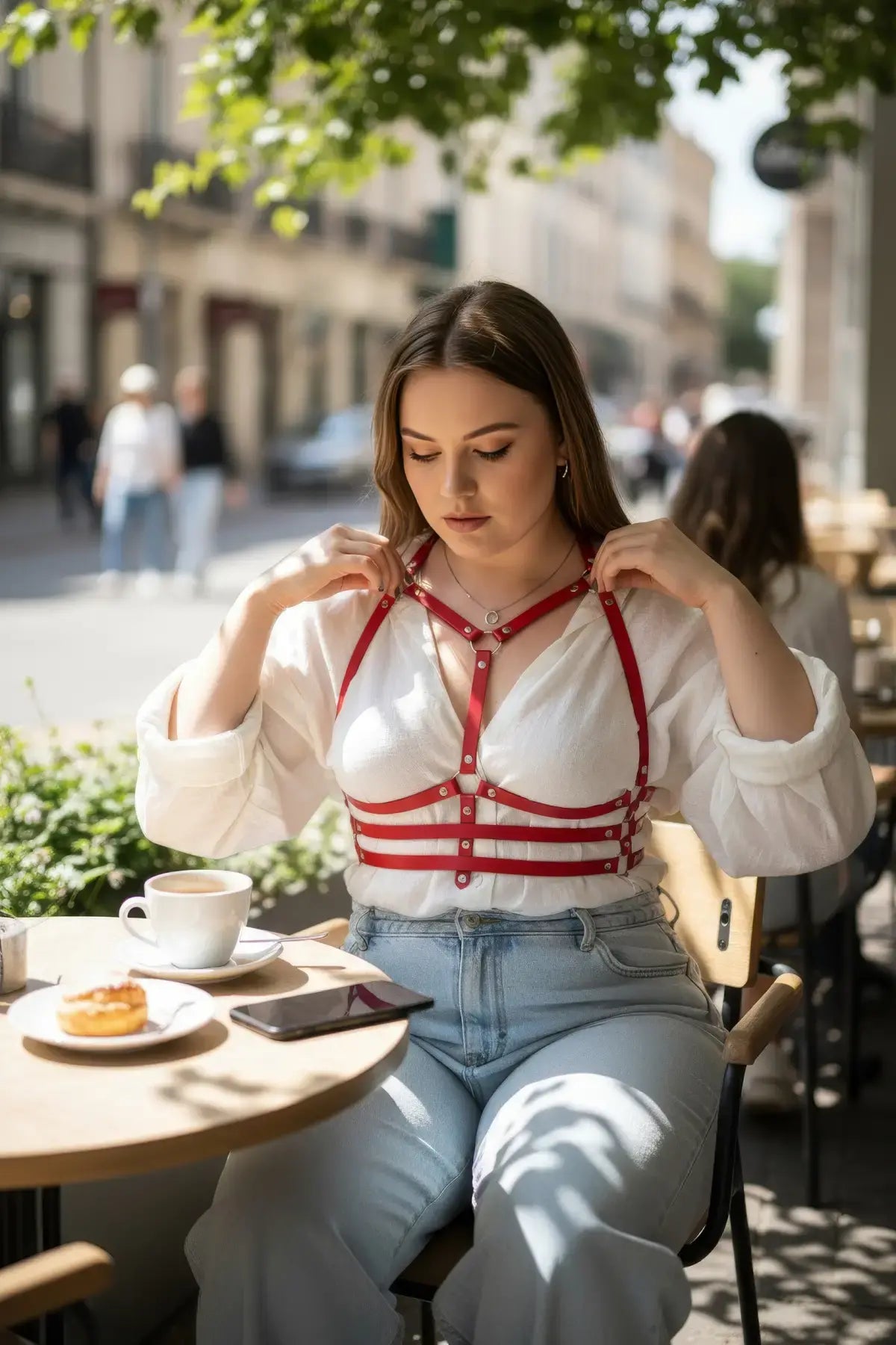 Plus-size red vegan-leather shoulder-strap corset harness worn over a white blouse at an outdoor cafe