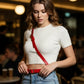 Red thin cross-body strap harness belt by HarnessClub, vegan leather worn over white ribbed top and blue jeans.